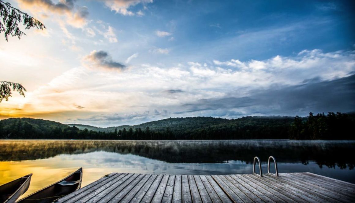 image of a morning scene over a cottage lake with dock and canoe in foreground and rising sun in the back lighting up the distance hills and forest