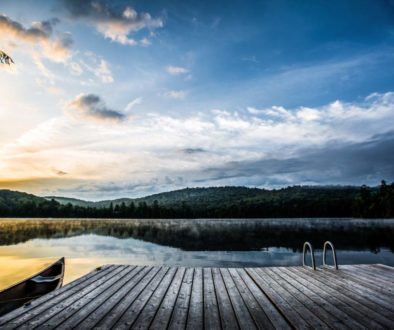 image of a morning scene over a cottage lake with dock and canoe in foreground and rising sun in the back lighting up the distance hills and forest