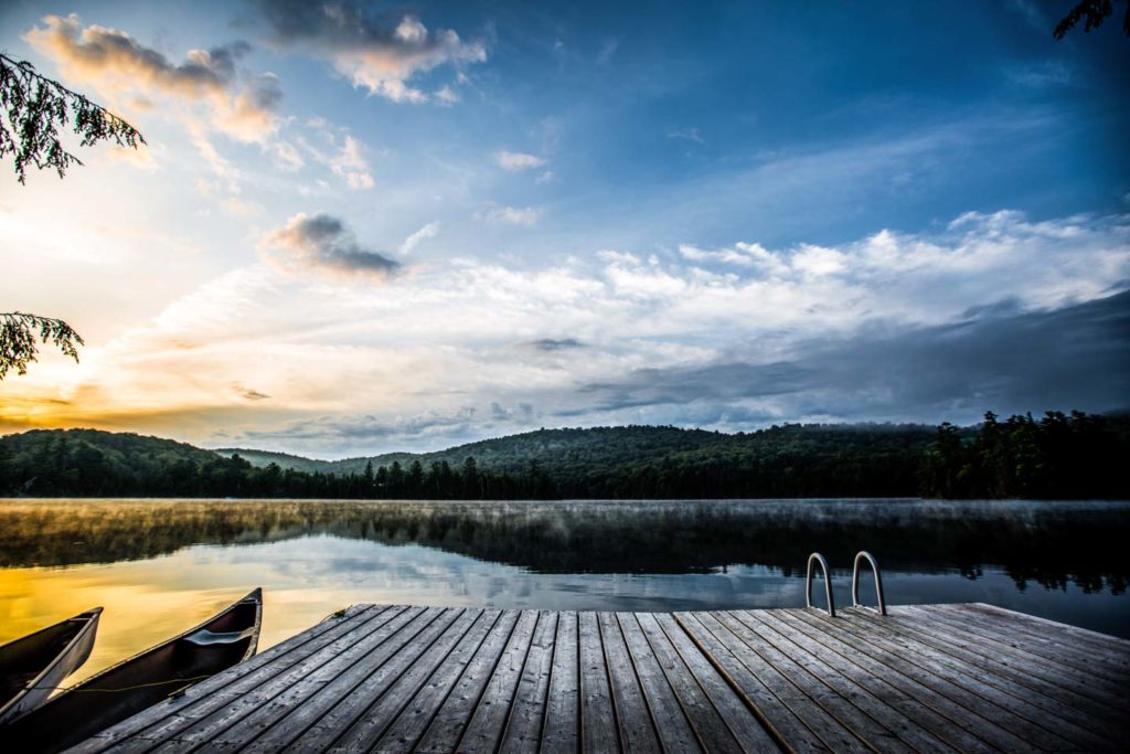 image of a morning scene over a cottage lake with dock and canoe in foreground and rising sun in the back lighting up the distance hills and forest